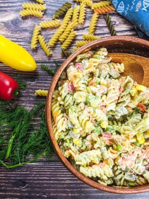 Fusilli pasta salad served in wooden bowl next to fresh dill, peppers and tricolore fusilli pasta.