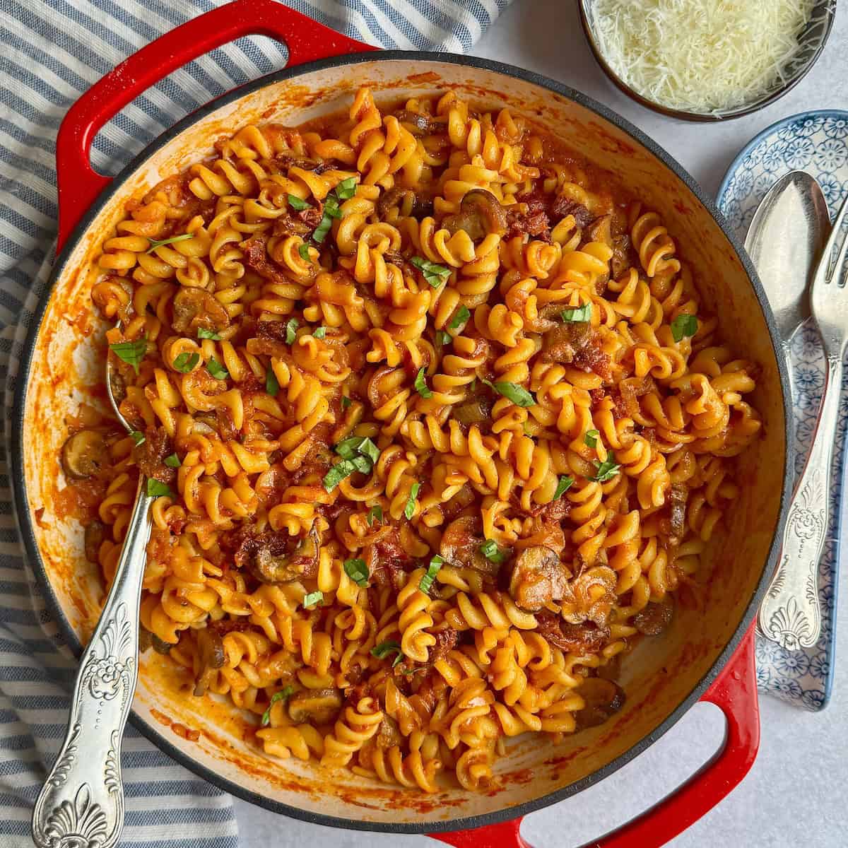 Pasta with sun-dried tomatoes and mushrooms in a red cast iron pan with chopped basil on top.