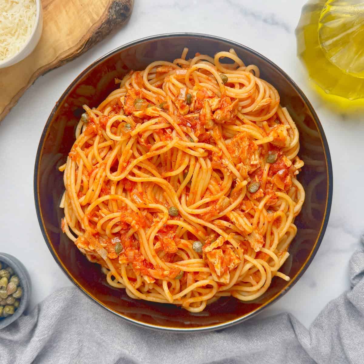 Served pasta with tuna in capers in black bowl with grated parmesan and bowl with capers next to it.