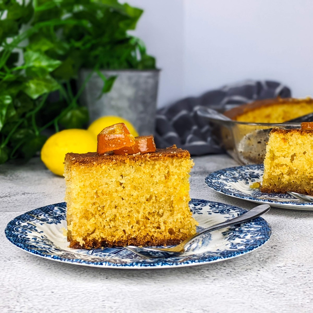Slice of ravani cake on a plate with lemons and tray of ravani at the back.