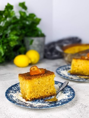 Slice of ravani cake on a plate with lemons and tray of ravani at the back.