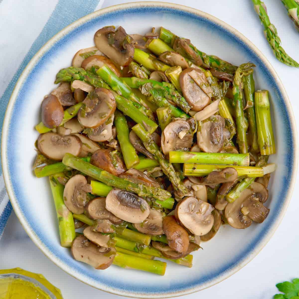Sauteed asparagus and mushrooms on a white bowl next to parsley, olive oil and asparagus.