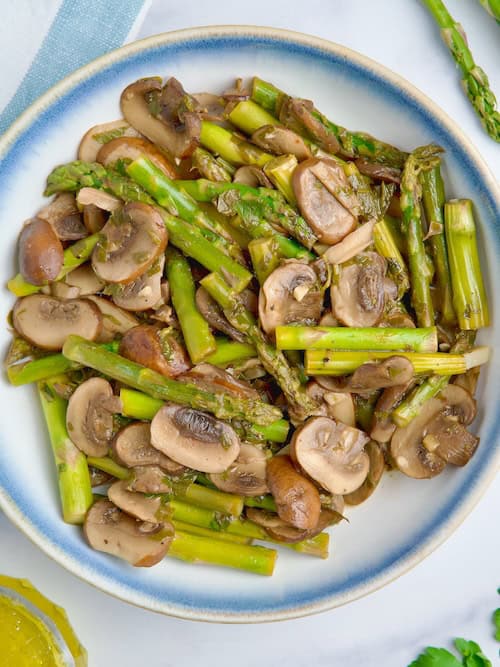 Sauteed asparagus and mushrooms on a white bowl next to parsley, olive oil and asparagus.
