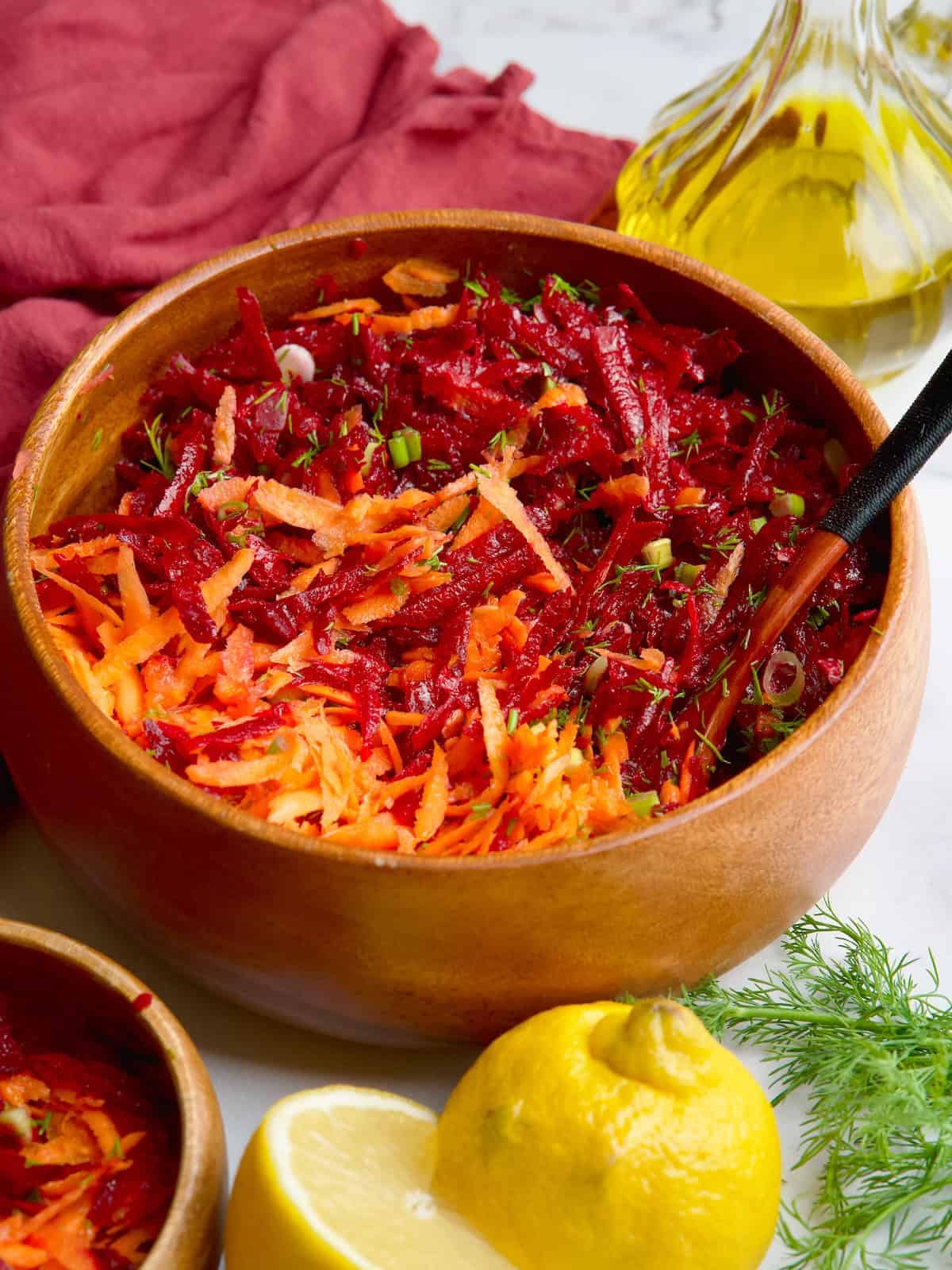 Beet and carrot salad in wooden bowl next to lemon slices, olive oil and fresh dill.