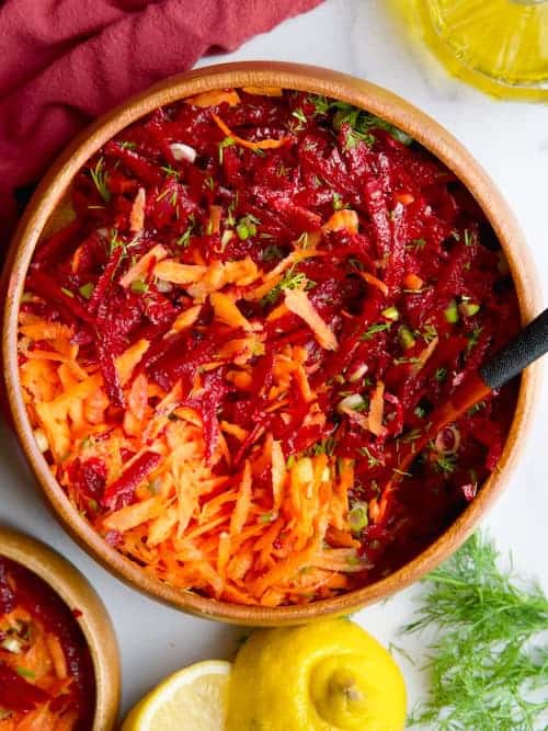 Beet and carrot salad in wooden bowl next to lemon slices, olive oil and fresh dill.