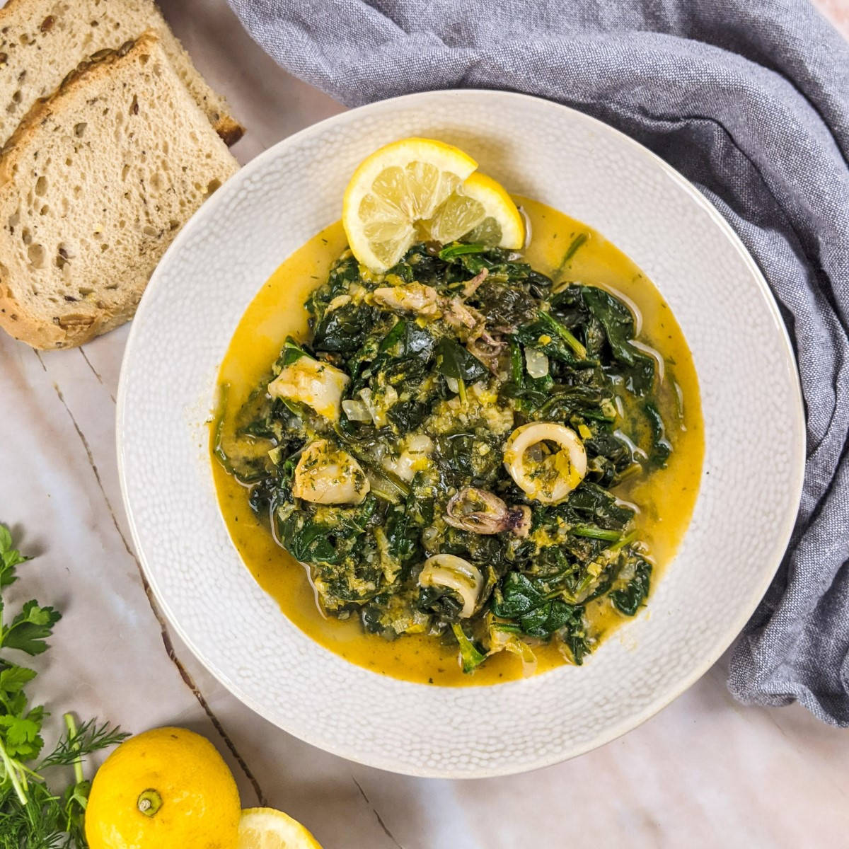 Squid stew with spinach served in a white bowl next to a lemon and some fresh dill and parsley.
