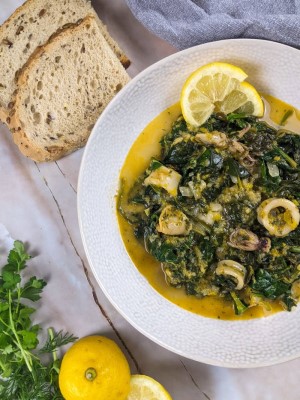 Squid stew with spinach served in a white bowl next to a lemon and some fresh dill and parsley.