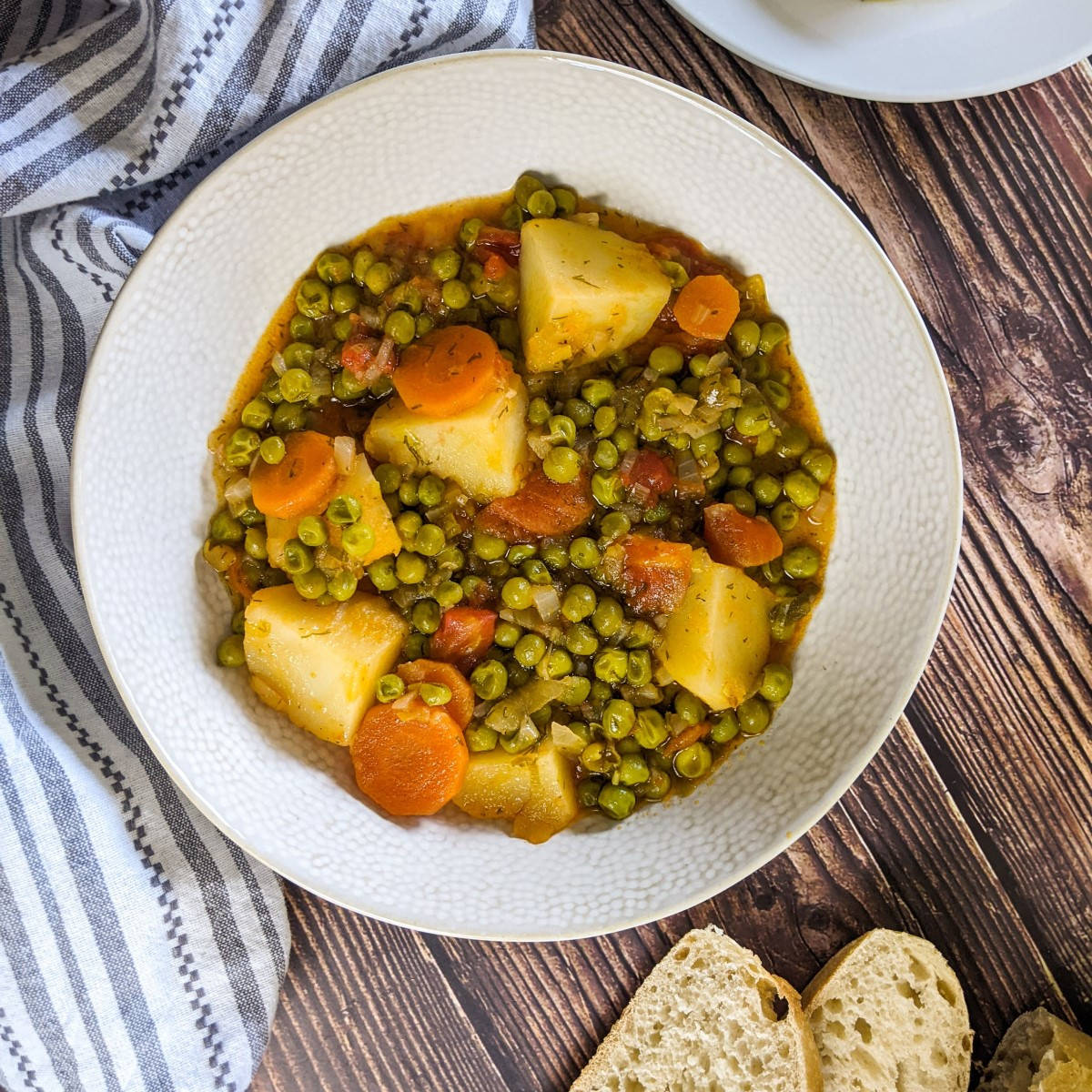 Stewed green peas served in a white bowl next to bread and feta cheese.