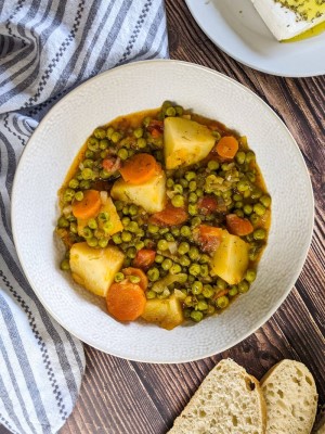 Stewed green peas served in a white bowl next to bread and feta cheese.