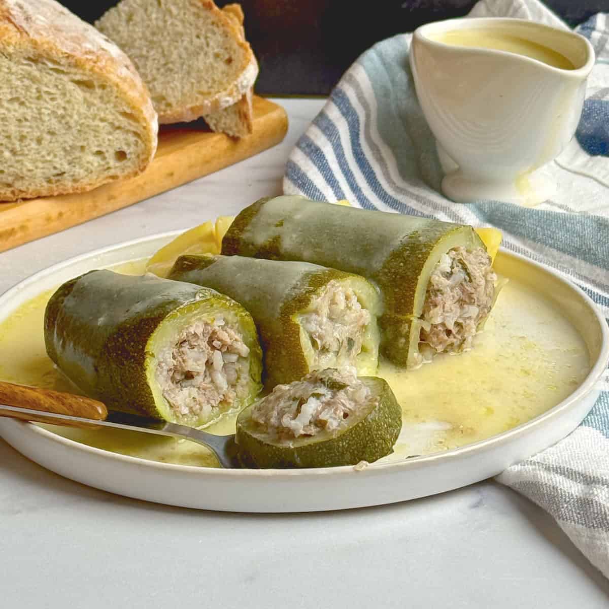 Stuffed zucchini in a white plate with egg lemon sauce poured on top and bread loaf at the back.