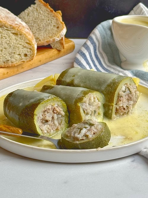 Stuffed zucchini in a white plate with egg lemon sauce poured on top and bread loaf at the back.