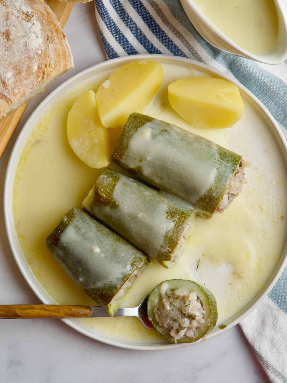 Stuffed zucchini in a white plate with egg lemon sauce poured on top and bread loaf at the back.