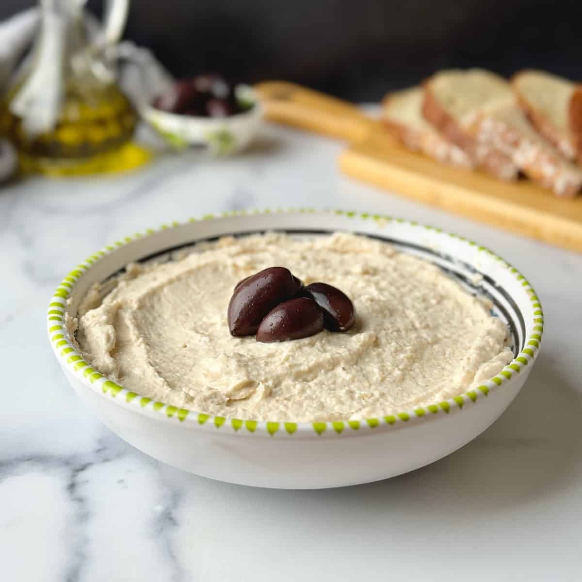 Taramosalata served in bowl plate with olives in the middles and bread slices at the back.