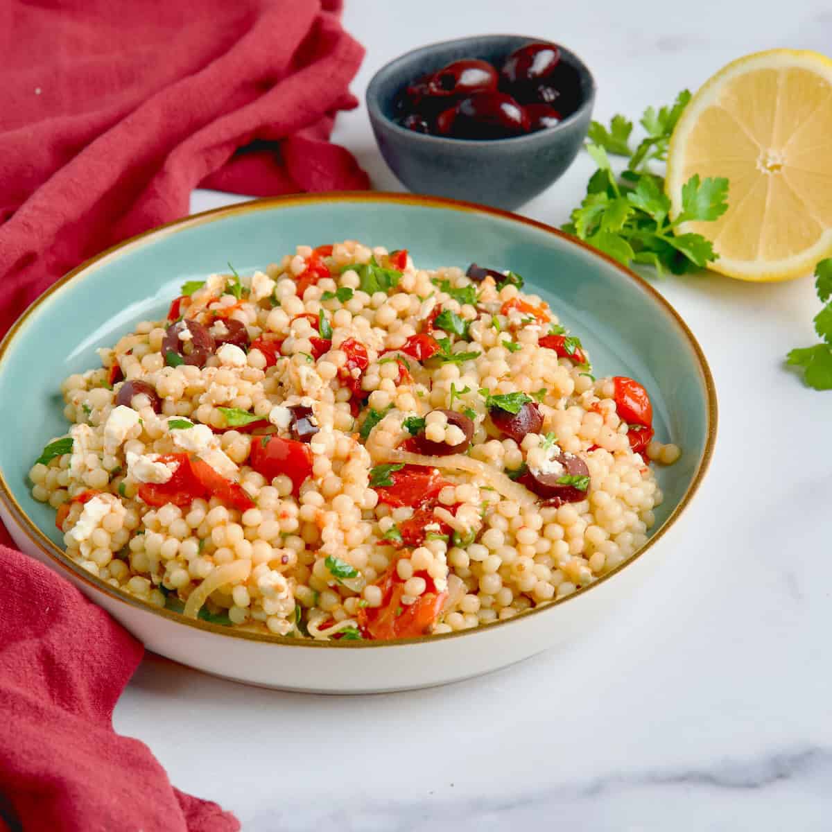 Pearl couscous with roasted tomatoes and olives salad served in a blue plate next to olives, lemon slices and parsley.