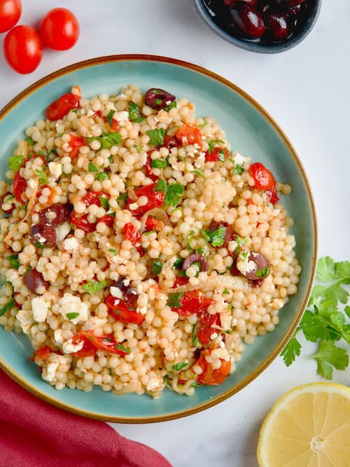 Pearl couscous with roasted tomatoes and olives salad served in a blue plate next to olives, lemon slices and parsley.