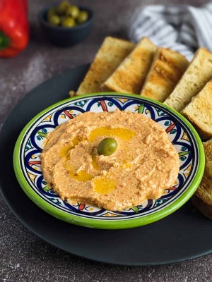 Greek spicy feta cheese dip served on a plate with an olive on top and next to slices of bread.